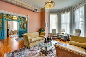Een ruime woonkamer met houten vloeren, een grote kroonluchter en klassiek meubilair, waaronder een bank, fauteuils en een glazen salontafel.
