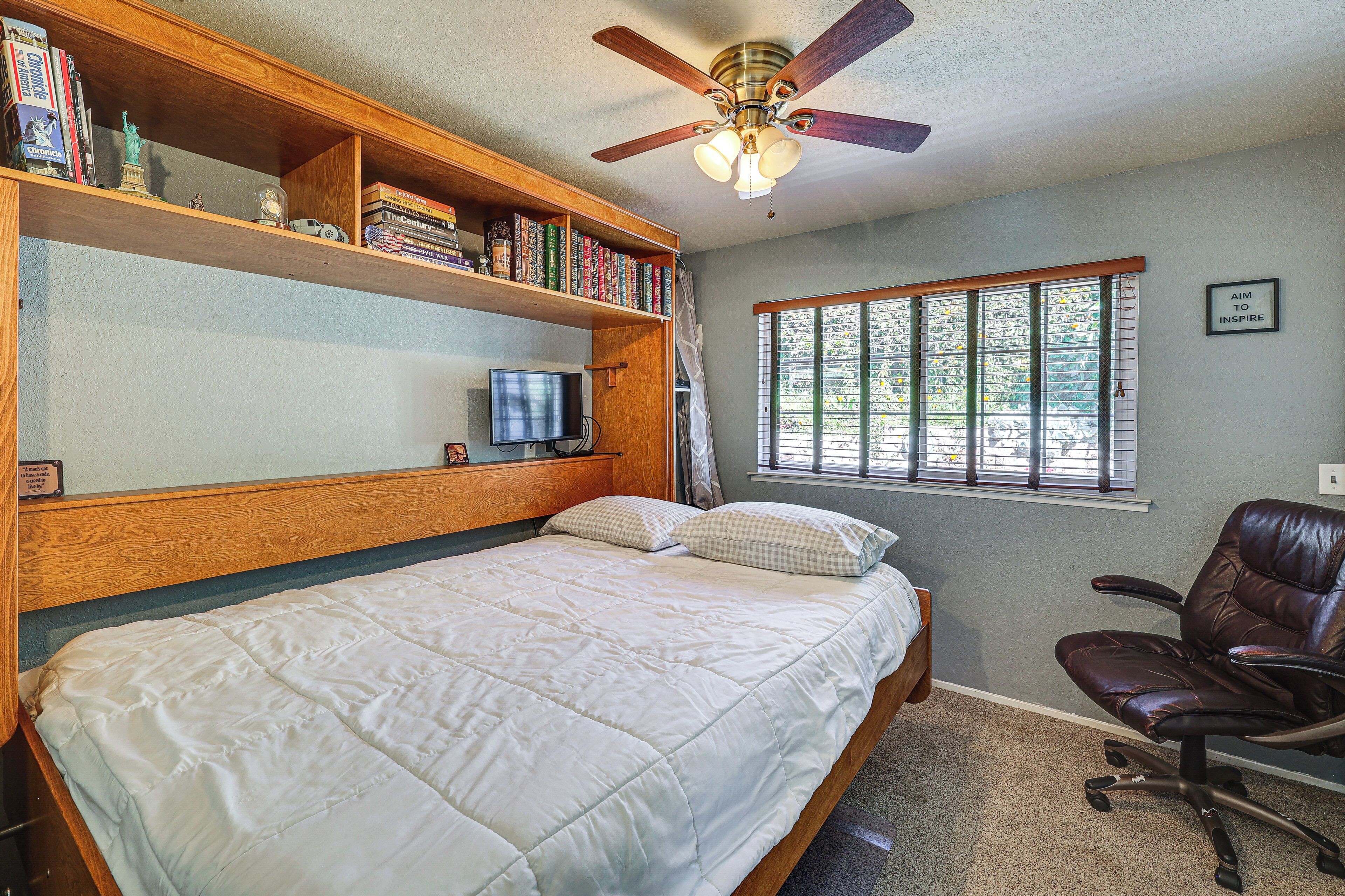 Un dormitorio con cama, escritorio con computadora, estantería, ventana con persianas y ventilador de techo.