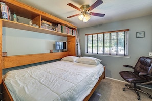 Ein Schlafzimmer mit einem Bett, einem Schreibtisch mit Computer, einem Bücherregal, einem Fenster mit Jalousien und einem Deckenventilator.