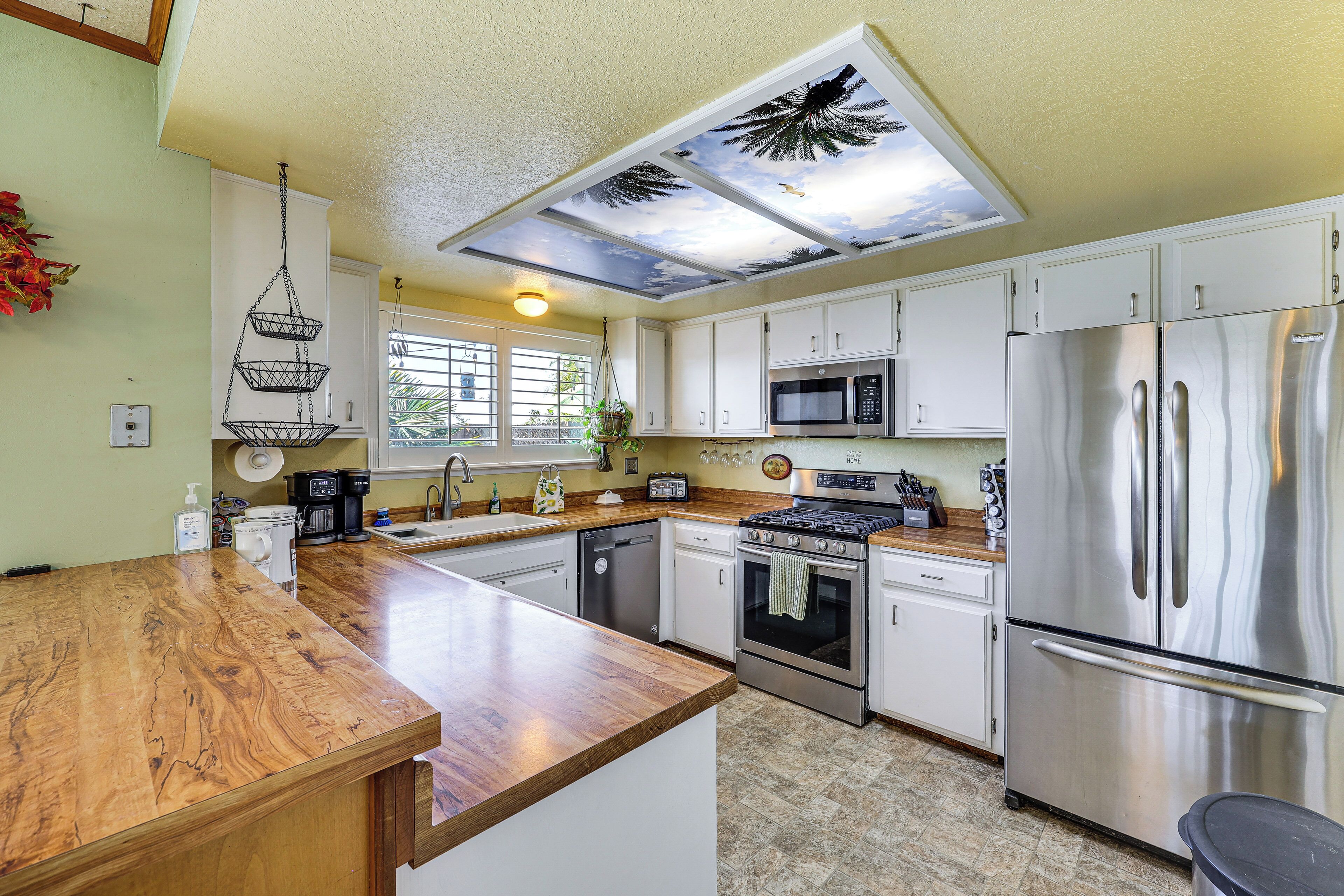 A kitchen with white cabinets, stainless steel appliances, a wooden countertop, and a ceiling with a tropical mural.