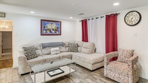 A living room with a beige sectional sofa, a glass coffee table, a patterned armchair, a clock, and a framed picture on the wall. - Meech Legacy House Southern Charm & Cozy Comfort (Benton)