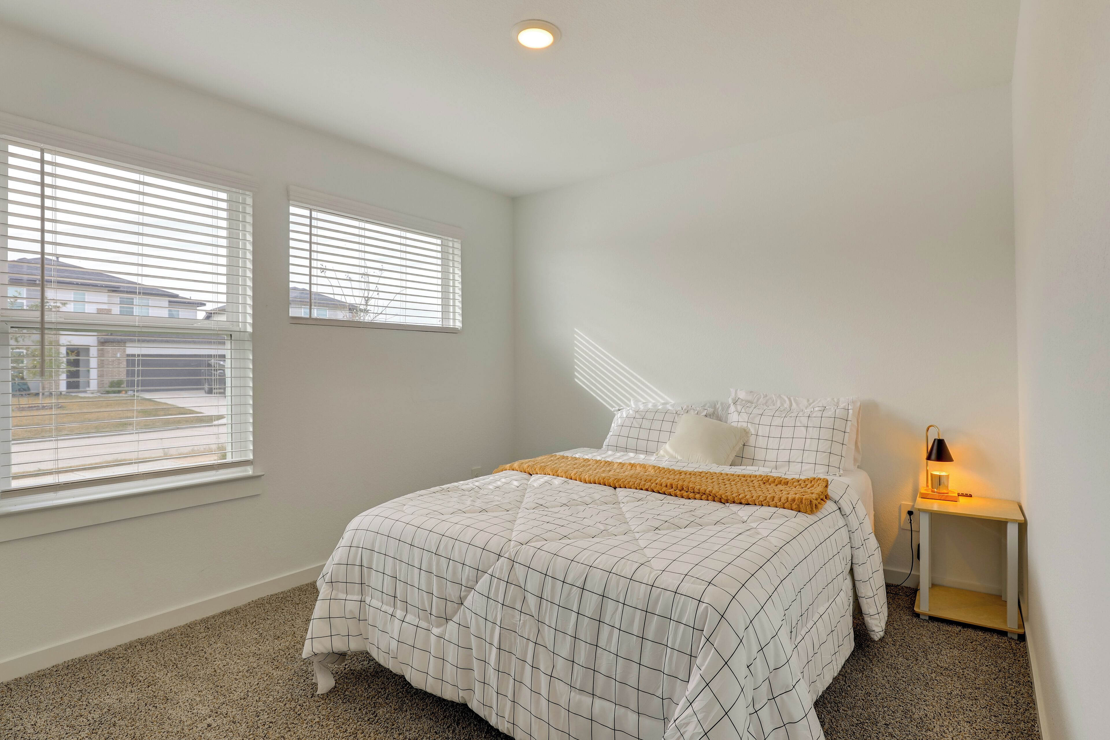 A bedroom with a bed, a nightstand, a lamp, and two windows with blinds.