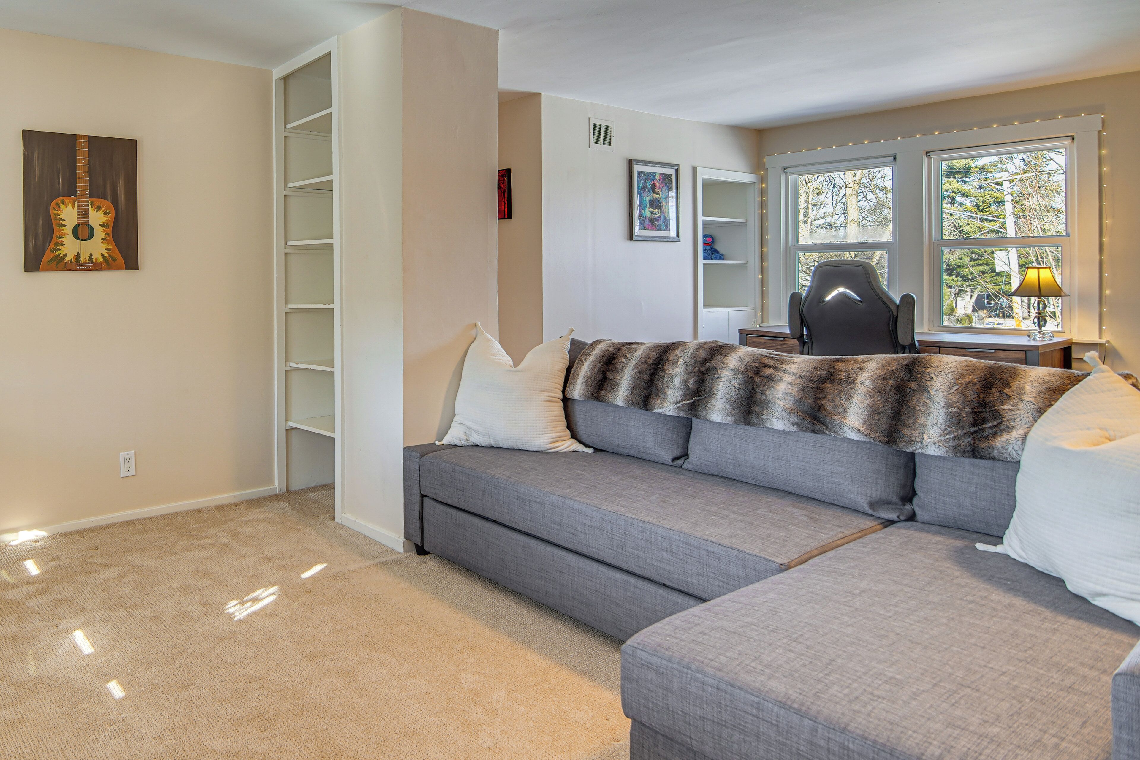 A living room with a grey sofa, a fur throw, and a chair. There are shelves and a window with a view.