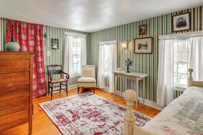 Een kamer met gestreept behang, een houten dressoir, een stoel, een bed en een tafel met een lamp.