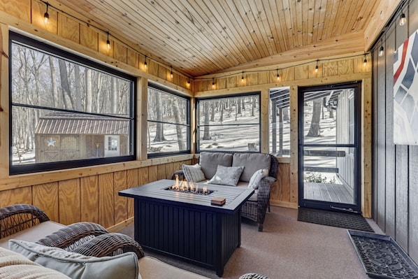 A cozy cabin interior with large windows, a central seating area with a fire feature, and a wooden ceiling.