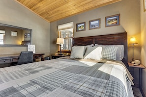 A bedroom with a large bed, a desk, a chair, a nightstand with a lamp, and a mirror.