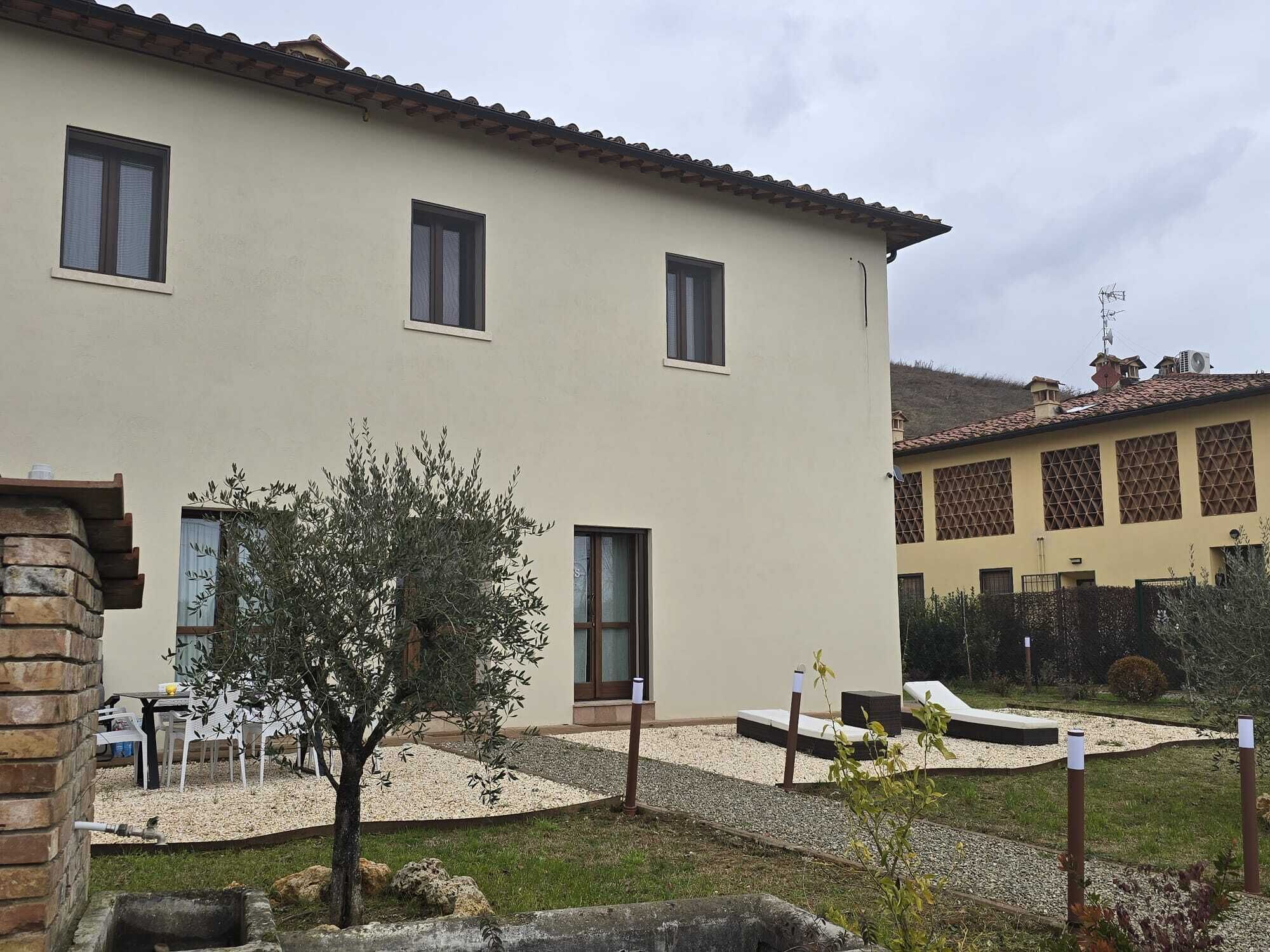 A two-story building with a beige exterior, multiple windows, and a small garden area with a tree and gravel paths.