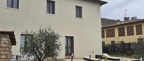 A two-story building with a beige exterior, multiple windows, and a small garden area with a tree and gravel paths.