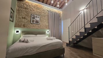 Un dormitorio con pared de piedra, cama con sábanas blancas, una escalera y una lámpara en el techo.