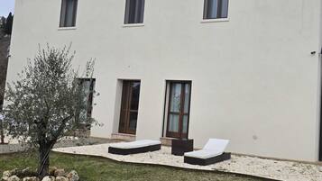 Una casa moderna de dos pisos con un patio que incluye dos tumbonas y un árbol pequeño.