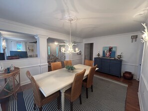 A dining area with a white table, wicker chairs, and a chandelier. There is a blue cabinet with decorative items on top. - In-town Lewes house with great floor plan!  112 Bradley Lane (Lewes)
