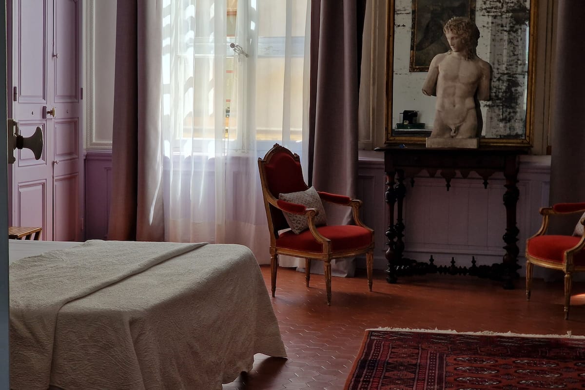 Una habitación con una cama, dos sillas rojas, una mesa con una estatua y un amplio ventanal con cortinas.