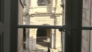 Une vue à travers une fenêtre ornée d’une grille en métal, donnant sur une structure arquée soutenue par des colonnes, ce qui évoque un site historique ou architectural.