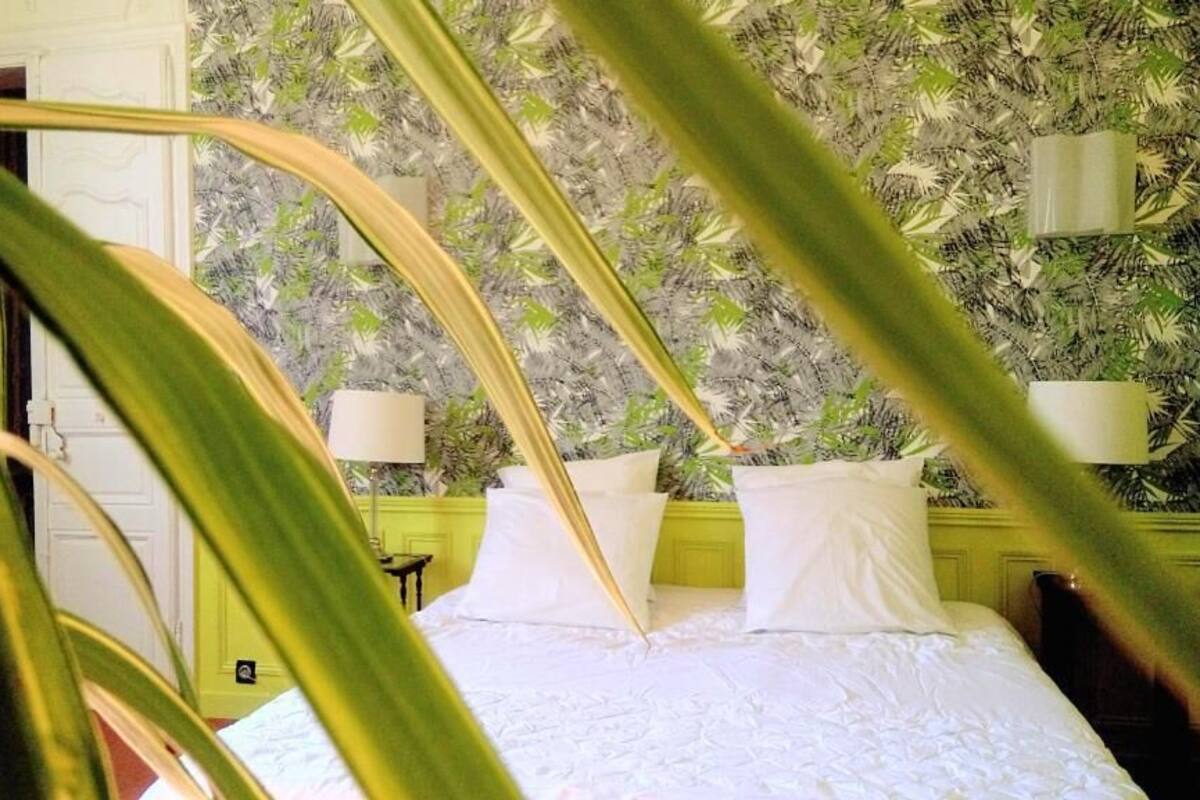 Un dormitorio con cama, ropa de cama blanca y pared verde con estampado de hojas.