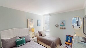 Une chambre avec deux lits, une table de chevet avec une lampe, une petite table avec une chaise, un téléviseur fixé au mur et des œuvres d’art.