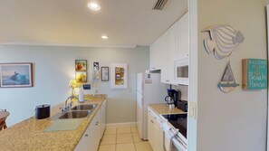 Une cuisine compacte équipée d’un évier, d’une cuisinière et d’un réfrigérateur. Les murs sont décorés, notamment avec un tableau représentant un phare et une pancarte de bienvenue.