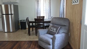 Espaço compacto para viver com cozinha compacta, área de refeições e um espaço de estar com uma poltrona cinza e um travesseiro com a inscrição 'STAY while'.