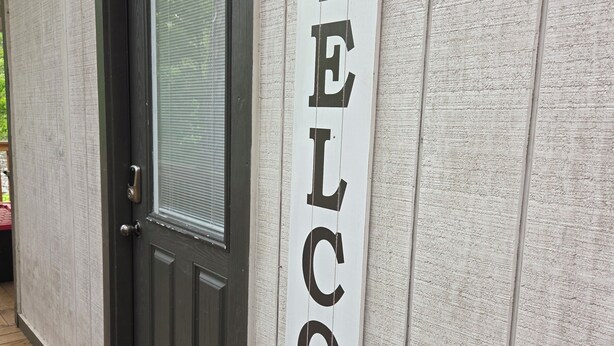 Uma porta de madeira com painel de vidro jateado e um toque de campainha preto. Uma placa vertical de boas-vindas está fixada na parede ao lado da porta.