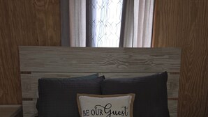 Cama com travesseiro que diz "BE OUR GUEST", uma toalha enrolada e cabeceira de madeira.