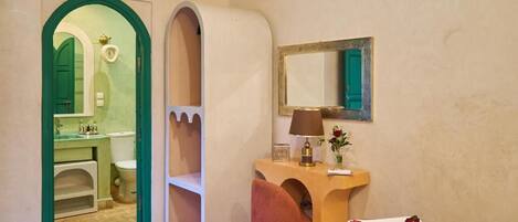 Une chambre d’hôtel avec un lit, une coiffeuse dotée d’un miroir, une porte à cadre vert donnant sur une salle de bain, et une table de chevet avec une lampe et des fleurs.
