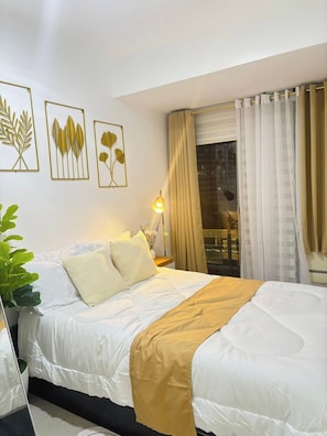 A hotel room with a bed, a bedside lamp, a window with curtains, and wall art. - Casa Filipina Subdivision (Parañaque)