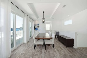 Ein geräumiger Essbereich mit einem großen Holztisch und weißen Stühlen, einer dunklen Holzbank an der Wand und großen Fenstern mit Blick nach draußen.