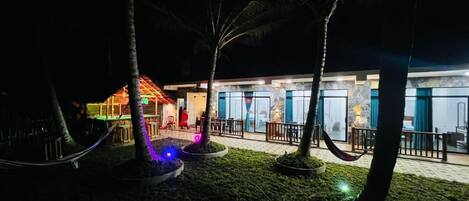 A nighttime view of a hotel with a well-lit outdoor area, including a pool and palm trees.