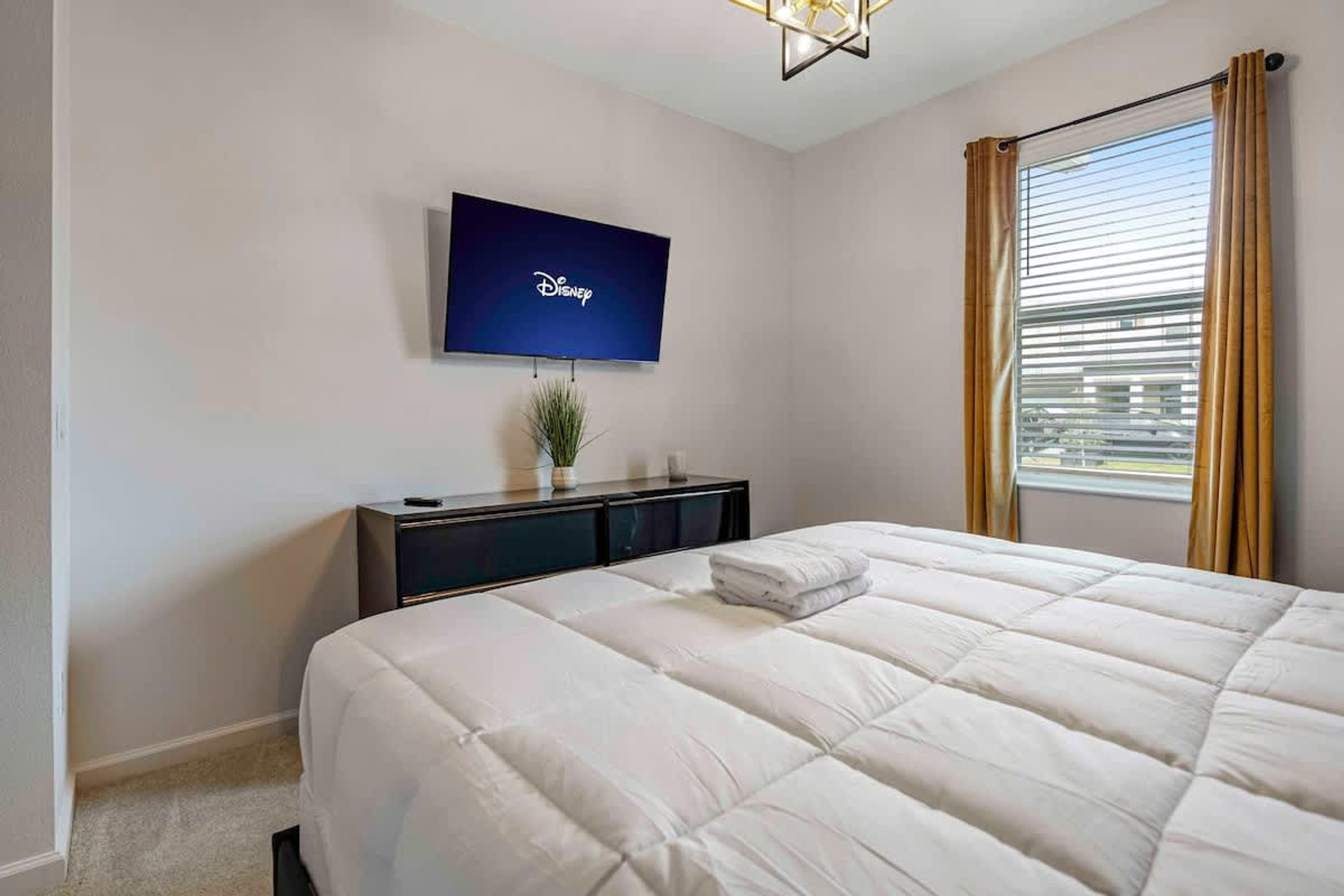A hotel room with a large bed, a flat-screen TV mounted on the wall, a bedside table with a plant, and a window with curtains.