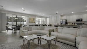 A modern living room with a sectional sofa, a coffee table, and a dining area with chairs and a table. - Magical 15br Villa w Pool Cinema Themed Fun (Kissimmee)