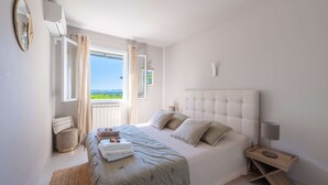 A bedroom with a large bed, a bedside table, a window with curtains, and a view of the outdoors. - Mas Noves - Horizon Alpilles Villa (Noves)