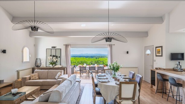 A spacious living area with a dining table set for six, a sofa, and a view of a vineyard through large windows. - Mas Noves - Horizon Alpilles Villa (Noves)