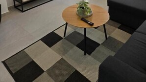 A living room with a flat-screen TV on a stand, a round coffee table with a plant, a black sofa, a checkered rug, and a door with a vertical handle. - Green Residence (Hersonissos)