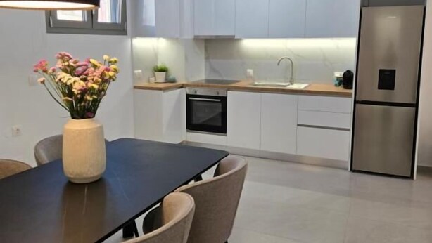 A modern kitchen with white cabinets, a stainless steel refrigerator, and a dining area with a dark table and chairs. - Green Residence (Hersonissos)