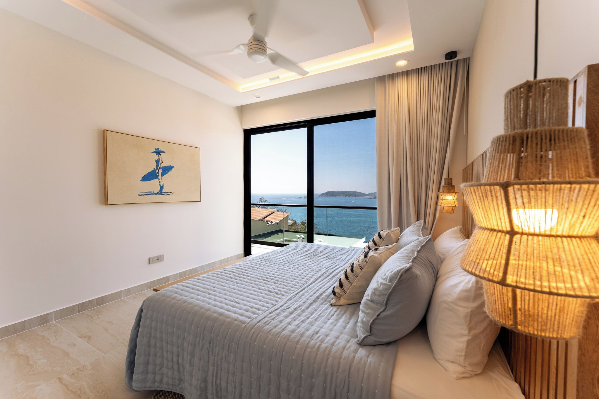 A modern bedroom with a large bed, a ceiling fan, a view of the sea, and a beachside balcony.