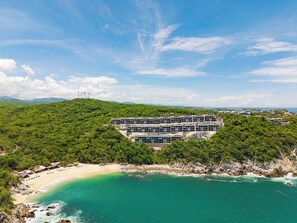 Ein Küstenhotel mit einem großen Gebäude, umgeben von üppiger Vegetation, einem Strand und klarem türkisfarbenem Wasser.