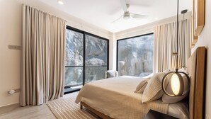 Een moderne slaapkamer met een groot bed, een plafondventilator en uitzicht op een rotsachtig landschap door het raam.