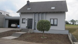 A modern two-story house with a grey tiled roof, a small front yard with a trimmed bush, and a garage with a white car inside.