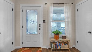 A white door with a glass panel, a window with blinds, and a small wooden console table with books and a plant. - Dean's Retreat At Georgetown 3 Bedroom Home (Georgetown)