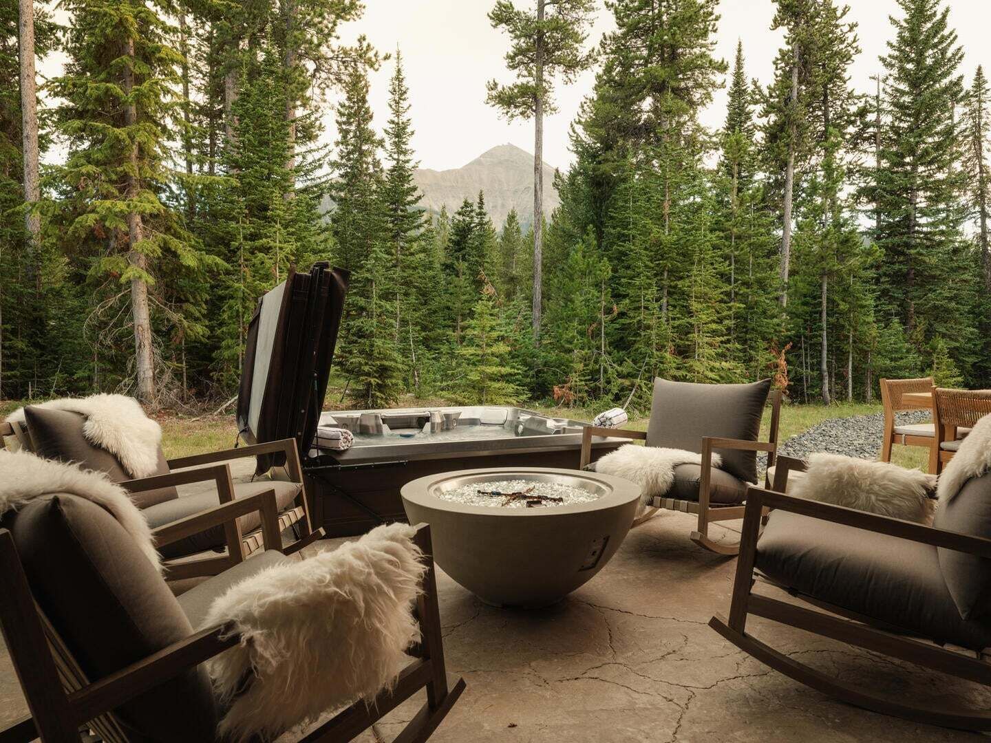 A cozy outdoor seating area with a fire pit, surrounded by tall trees and a mountain in the background.