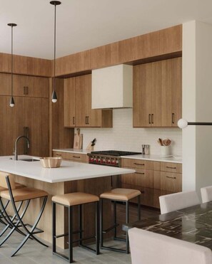 A modern kitchen with wooden cabinets, a central island with stools, pendant lighting, and a stainless steel sink. - 8 Speedwell 4 Bedroom Home (Big Sky)