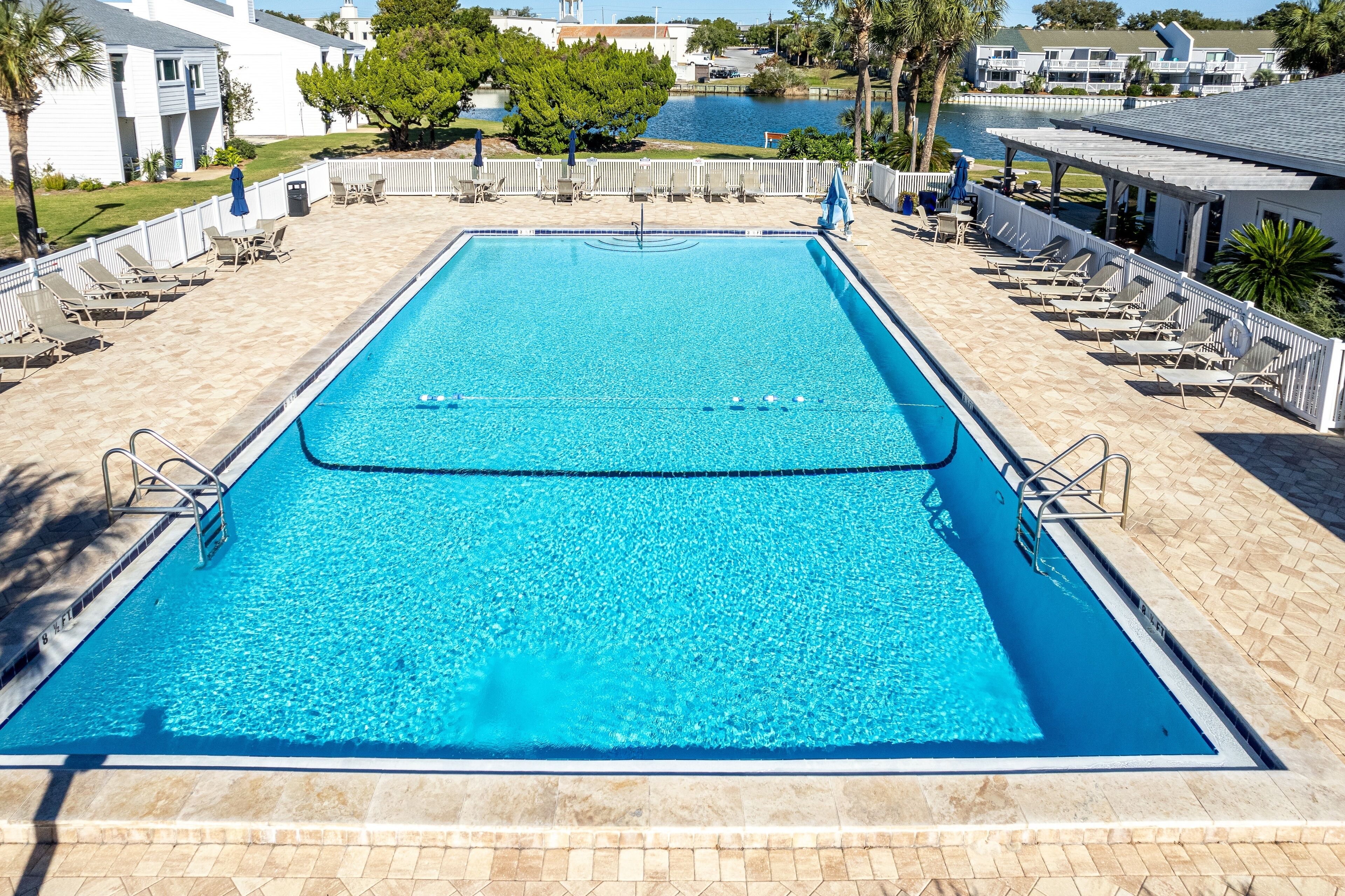Una piscina rectangular con agua azul cristalina, rodeada de sillas de descanso y sombrillas, con vistas a una masa de agua y edificios al fondo.
