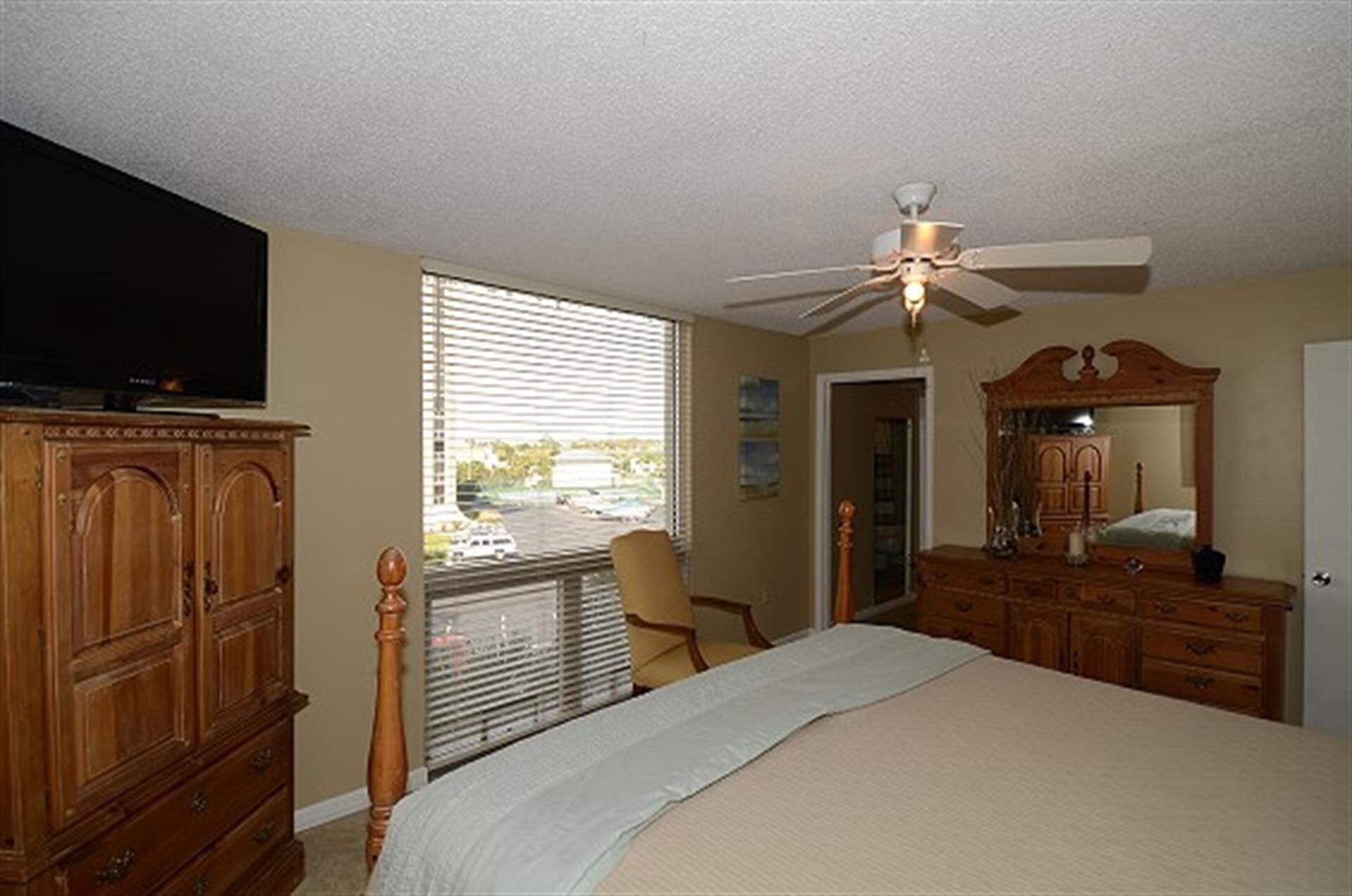 A bedroom with a bed, wooden dresser, television, ceiling fan, and a window with blinds.
