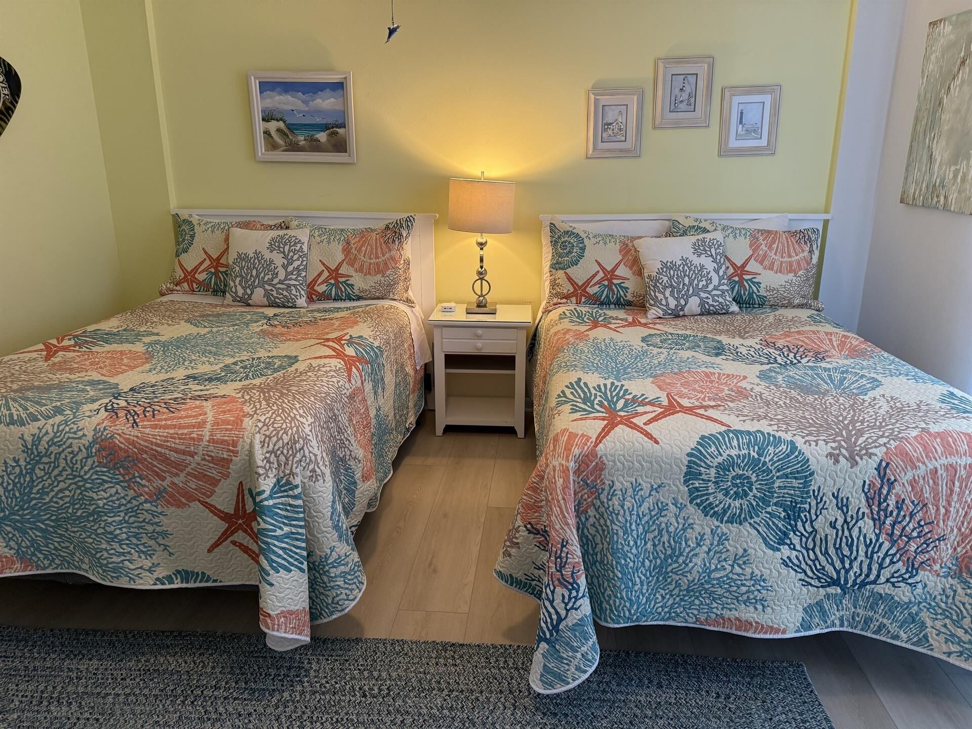 Une chambre avec deux lits, chacun agrémenté d’une couvre-lit aux motifs coralliens et d’étoiles de mer, une table de chevet avec une lampe et des tableaux encadrés accrochés au mur.