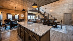 A spacious kitchen with granite countertops, wooden cabinets, and a central island. A staircase leads to an upper level, and there is a dining area with wooden tables and chairs. - Skywalk 5 Bedroom Home (Sevierville)