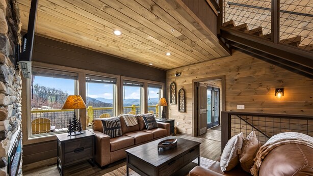 A cozy living room with a wooden ceiling, a sofa, a coffee table, and a view of the mountains through large windows. - Skywalk 5 Bedroom Home (Sevierville)