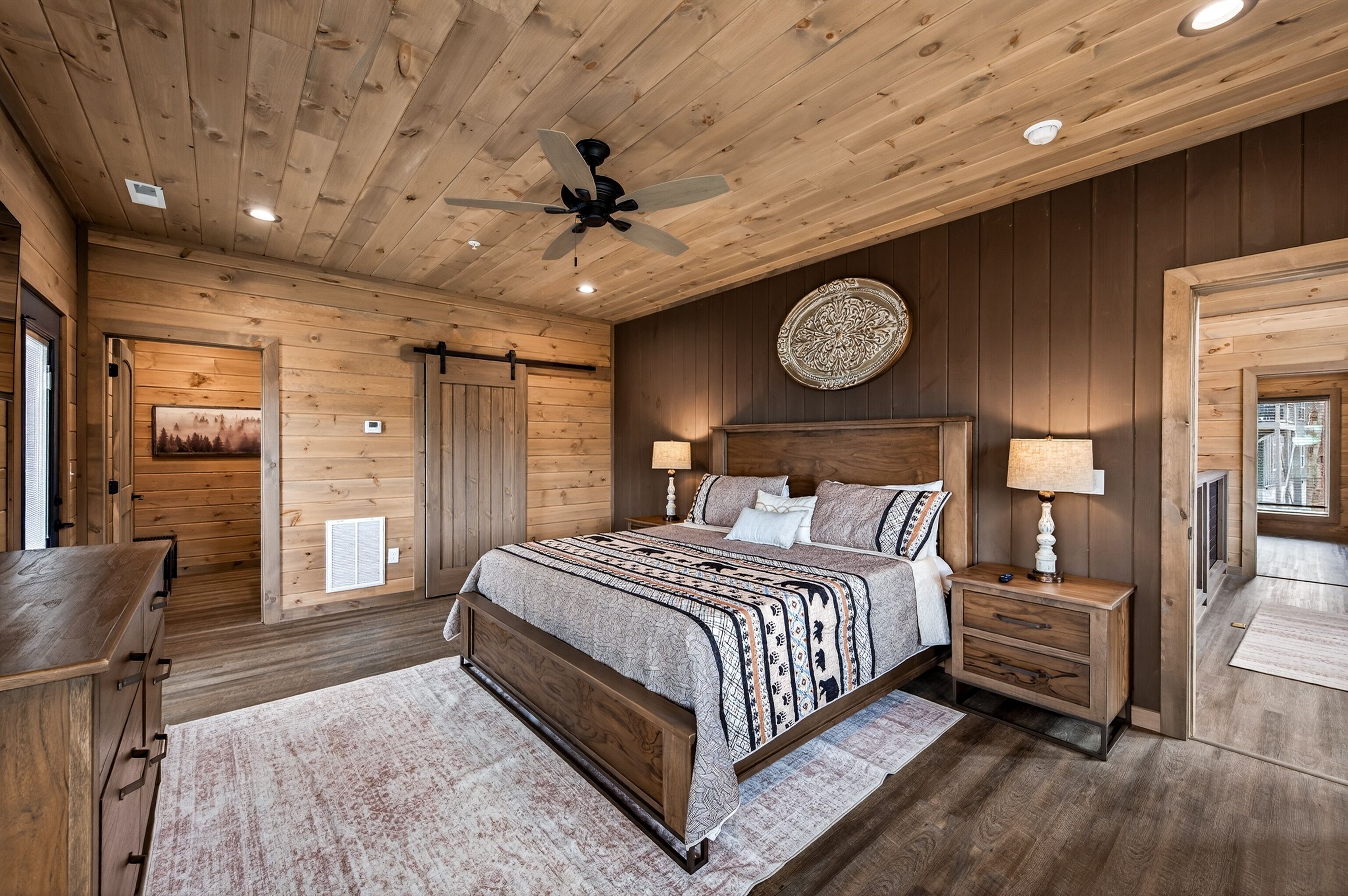 Un dormitorio con paneles de madera, una cama, mesitas de noche, un ventilador de techo y un elemento decorativo en la pared.