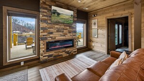 A cozy living room with a stone fireplace, wooden ceiling, and large windows overlooking a deck. - Skywalk 5 Bedroom Home (Sevierville)