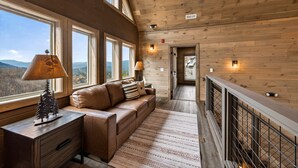 A cozy cabin interior with a wooden ceiling, large windows, a leather sofa, a nightstand, and a lamp. - Skywalk 5 Bedroom Home (Sevierville)