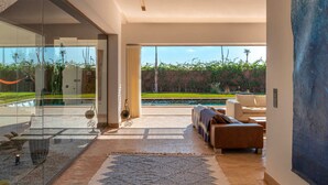 A modern living room with a large glass wall, a pool, and a view of greenery. - Villa Zen'O | Palmeraie view | Heated swimming pool (Marrakech)