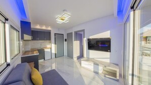 Ein modernes Wohnzimmer mit einer Couch, einem Couchtisch und einem Fernseher. Es gibt eine Küchenzeile mit Schränken und einer Arbeitsfläche. Das Zimmer hat große Fenster und einen Ausblick nach draußen.